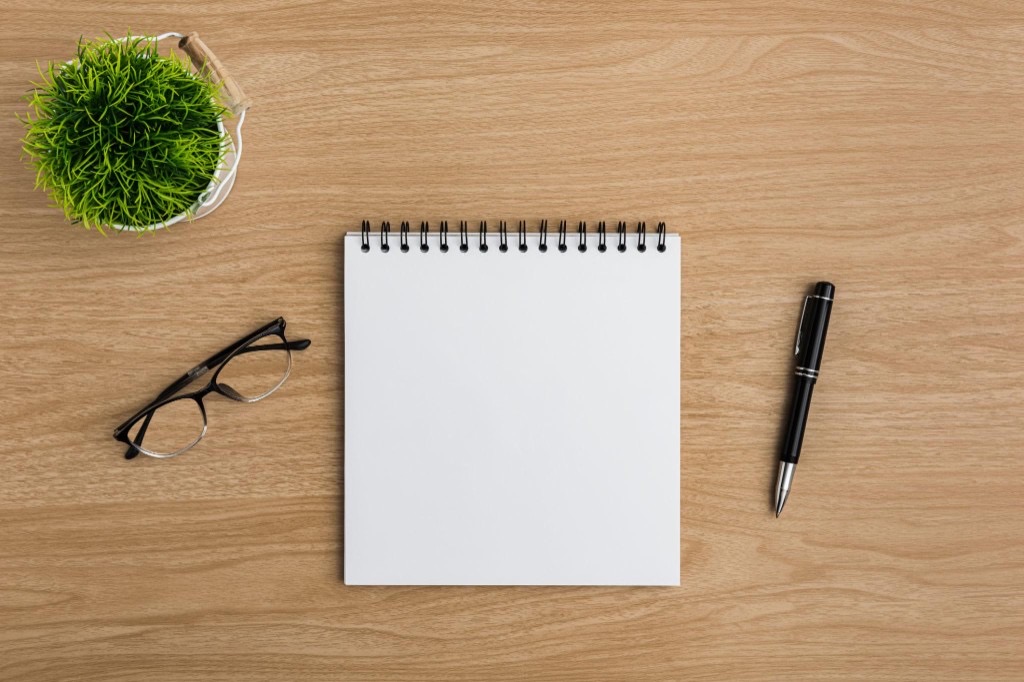 Top-down view of a blank spiral notebook on a wooden desk with glasses, a pen, and a small potted plant